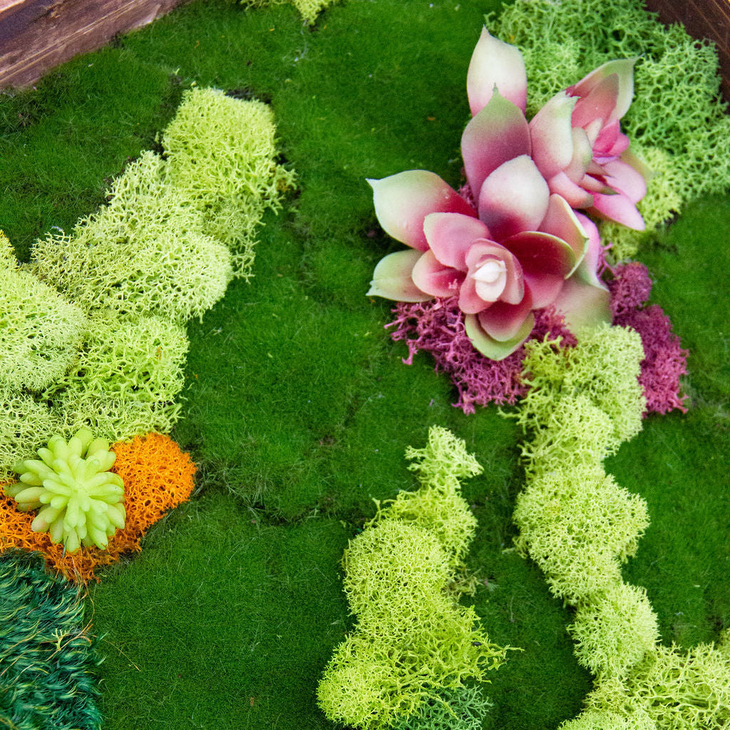 Decorative wreath made of green moss and pink artificial flowers on a wooden surface.