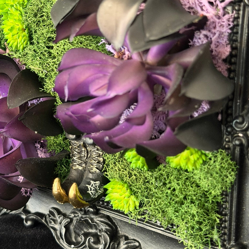 Decorative arrangement with purple flowers and greenery in an ornate frame.